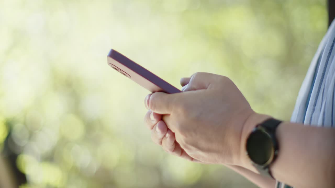 Anziano uomo asiatico che tiene in mano un telefono viola, toccando, scorrendo e aggiustando la presa all'esterno, indossando uno smartwatch.