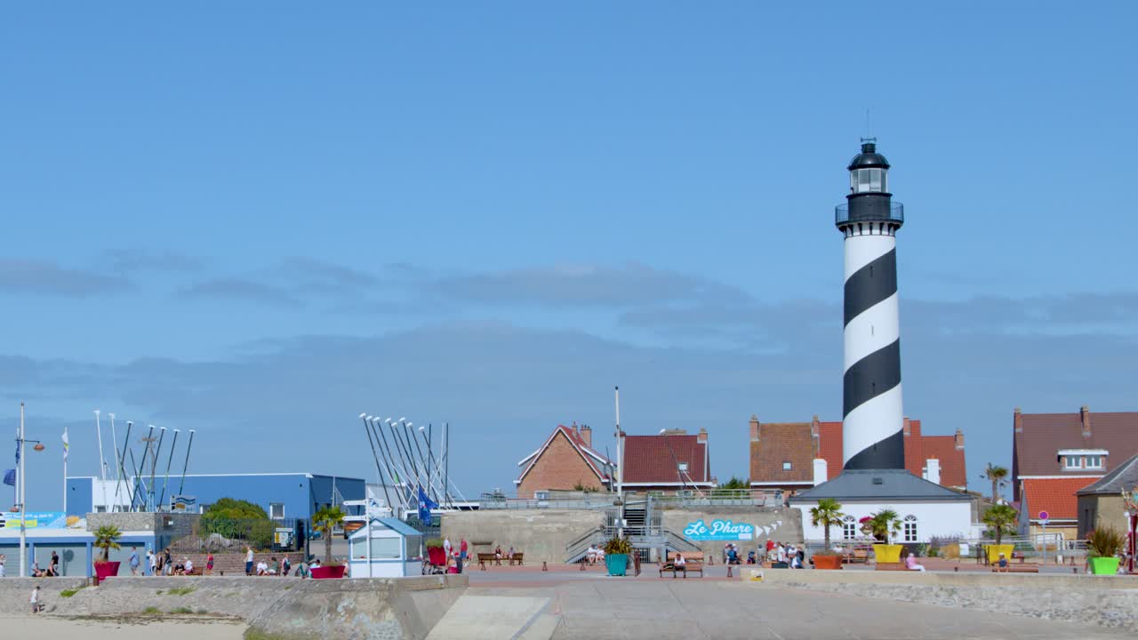 Daytime wide shot of striped lighthouse, beach, and town buildings under clear blue sky