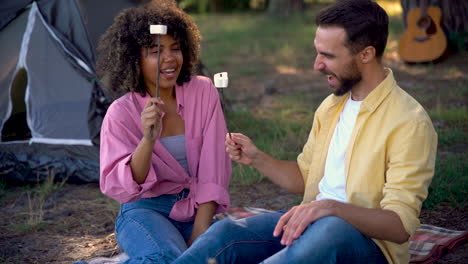 happy hiker couple ready to to roast marshmallows on a bonfire, having fun. caucasian man book and young black female camping in the forest.