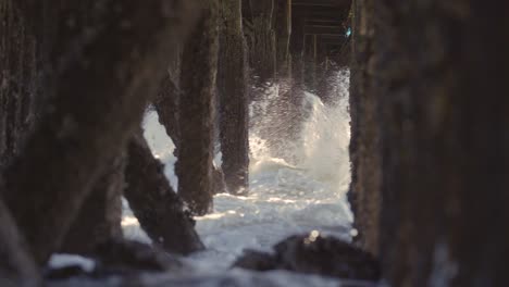 Waves-crashing-under-a-pier,-old-wooden-pillings-with-sunbeams-coming-through