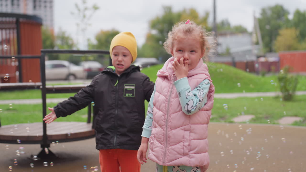 Playful boy in yellow hat bends forward trying to catch floating bubbles while girl in pink vest stands nearby smiling, surrounded by soft light, green park background, and joyful atmosphere