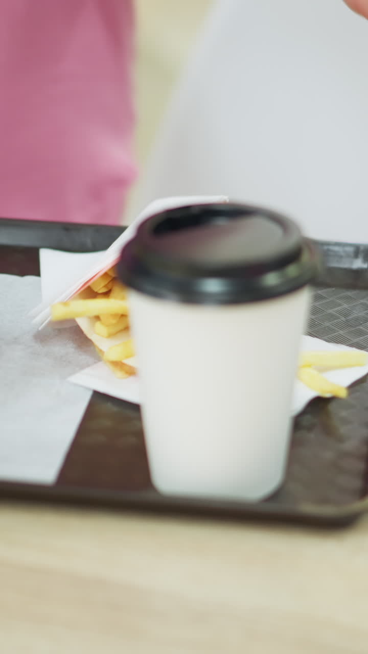 vista de la mano de una mujer en vestido rosa llevando una bandeja que contiene hamburguesa, papas fritas y café en una mesa de madera, se sienta y toma un chip de patata, con un fondo borroso del restaurante