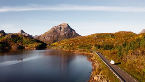 Volando-A-Través-De-Las-Hojas-Doradas-De-Los-árboles-Y-Sobre-Un-Lago,-Orbitando-Alrededor-De-Una-Camioneta-Blanca-Que-Se-Abre-Camino-En-La-Carretera,-Islas-Lofoten,-Noruega