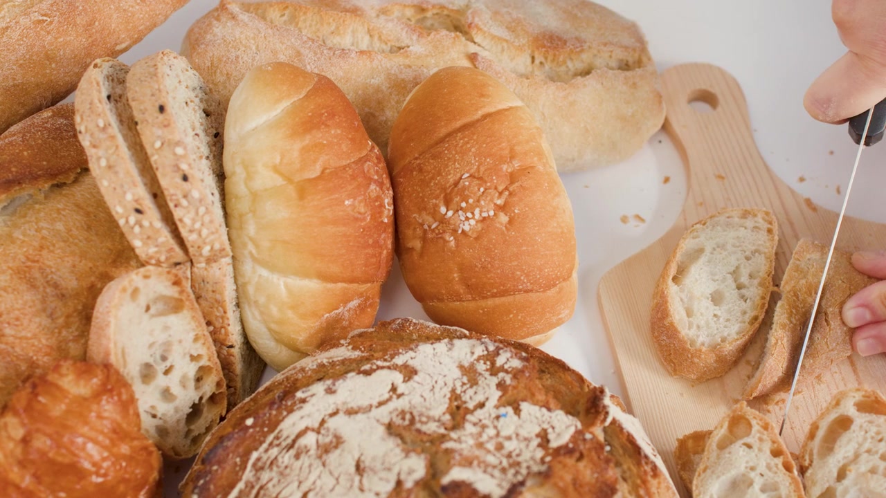 Close-up shot of hands slicing crusty bread on a board with assorted bakery loaves.