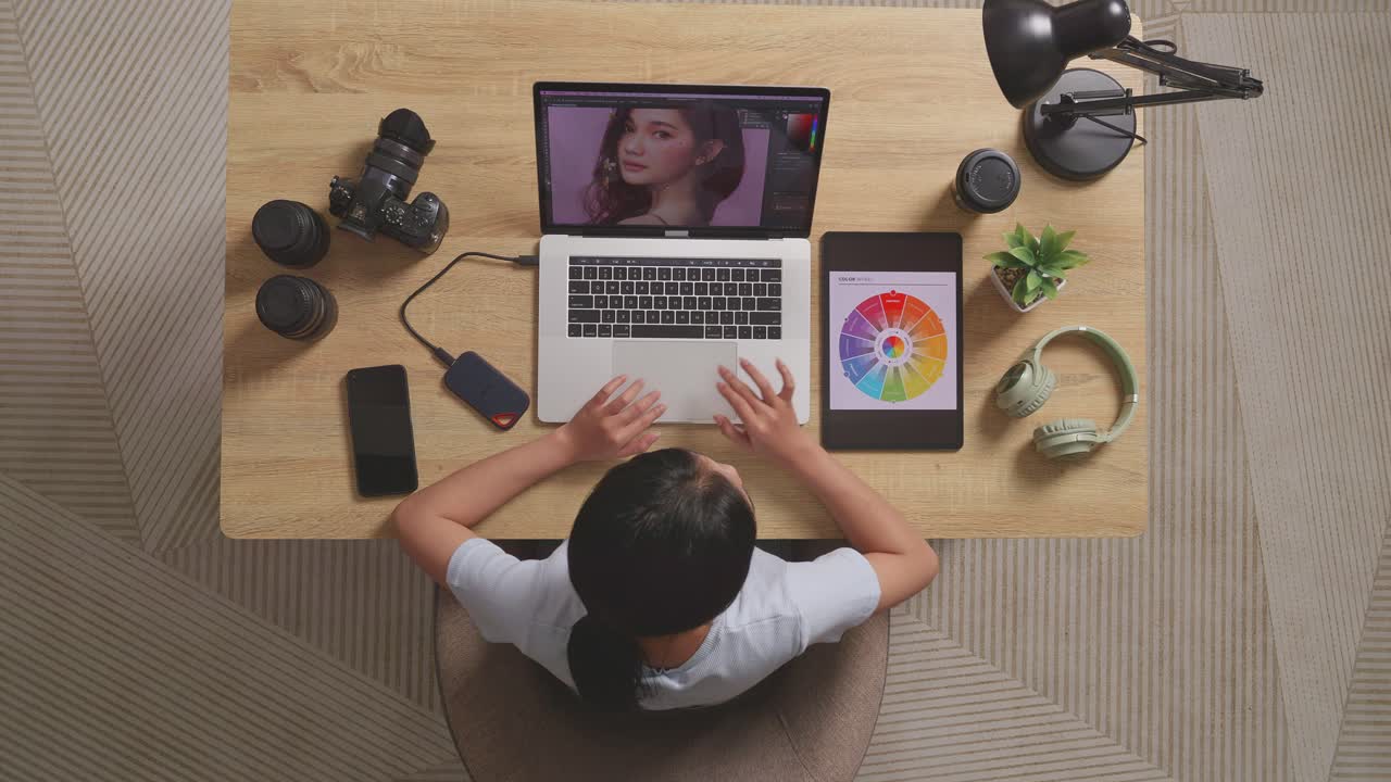 vista superior de una mujer editora mirando los esquemas de colores en una tableta mientras está sentada en el espacio de trabajo usando una computadora portátil al lado de la cámara editando la foto de una mujer en casa
