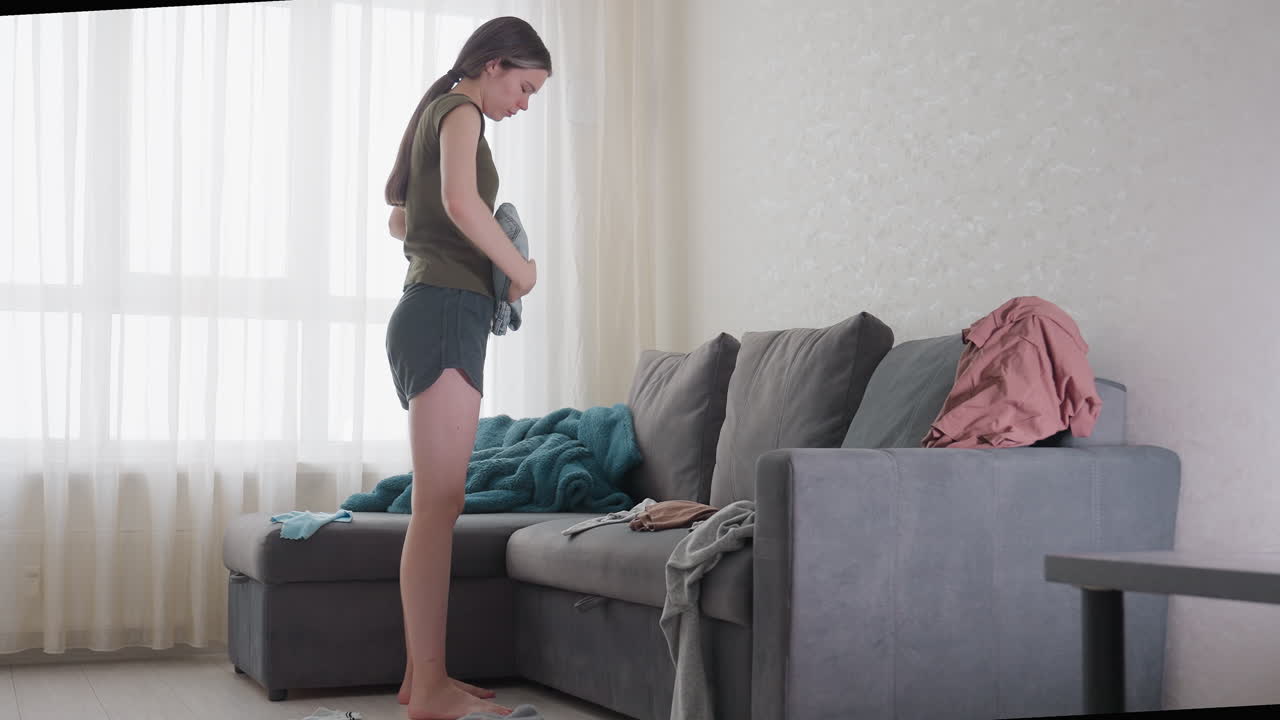 Young lady folding clothes neatly and placing them on grey sofa in bright living room, casual outfit in shorts and t-shirt, daylight streaming through window curtains