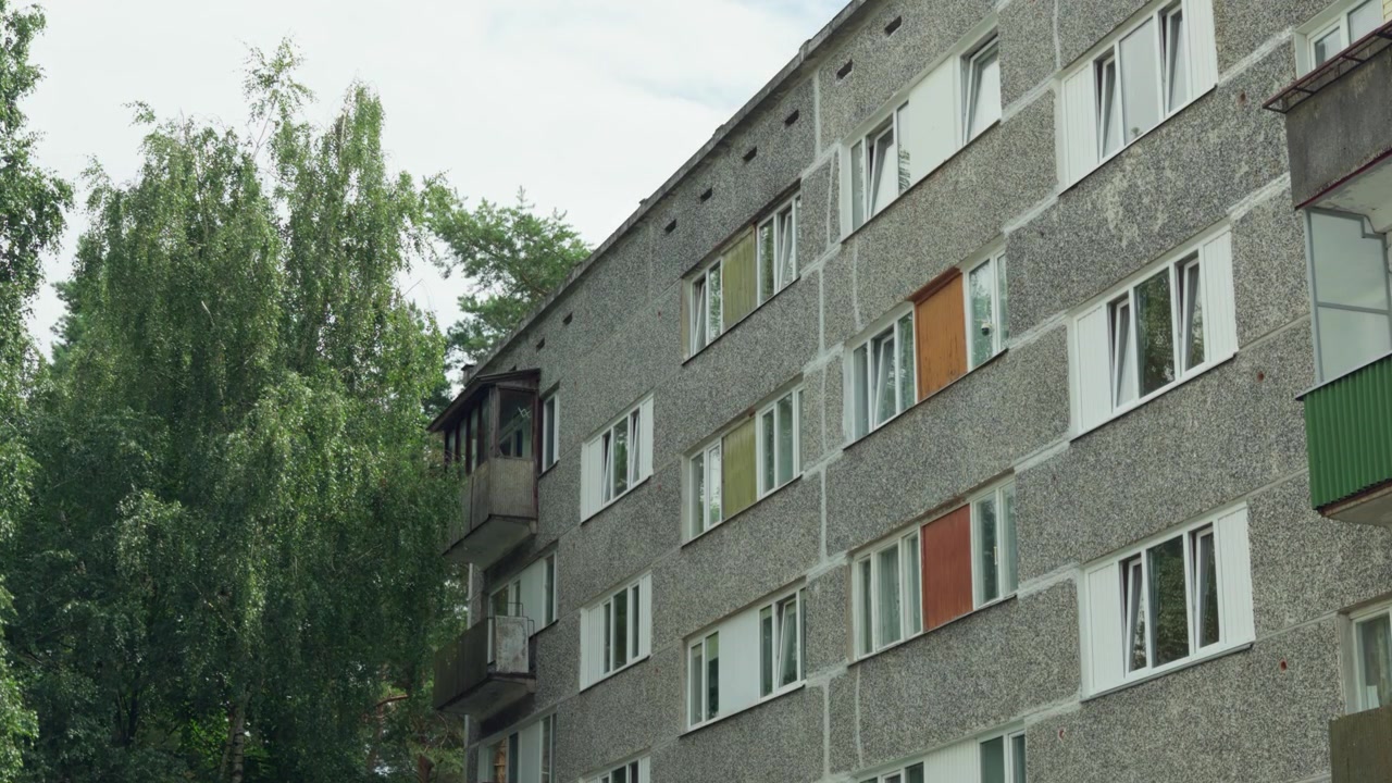 Bilde tatt nedenfra og opp som viser teksturert panelfasade av en boligblokk fra sovjettiden med fargerike balkonger ved siden av høye trær i Daugavpils