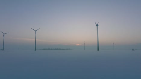 Aerial-view-of-wind-turbines-generating-renewable-energy-in-the-wind-farm,-snow-filled-countryside-landscape-with-fog,-sunny-winter-evening-with-golden-hour-light,-wide-drone-shot-moving-forward-low