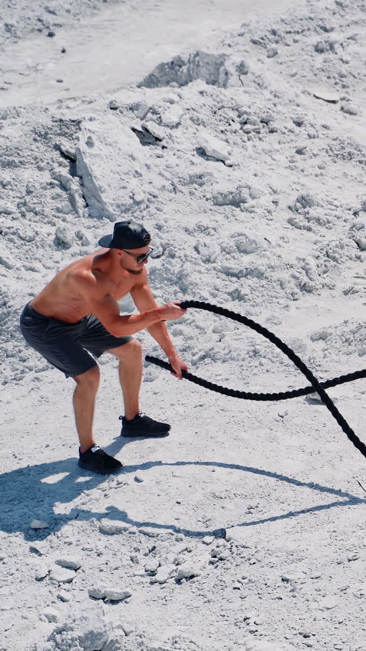 Athletic male performs battle ropes exercise on the white rocky background. Sportsman in shorts doing waves with cable during an outdoor fitness workout. Vertical video