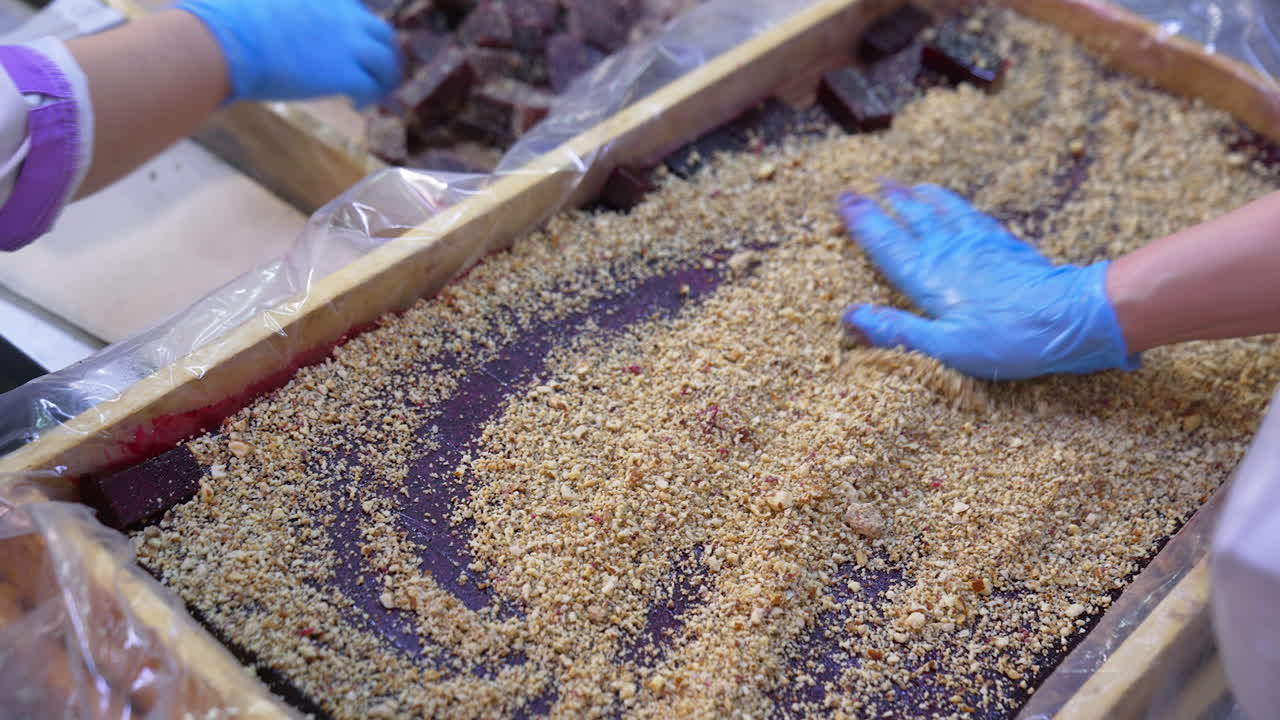 Purple dark layer of marmalade cut into pieces in a wooden box. Worker spreads nuts over the sweet candied fruit jelly. Close up.