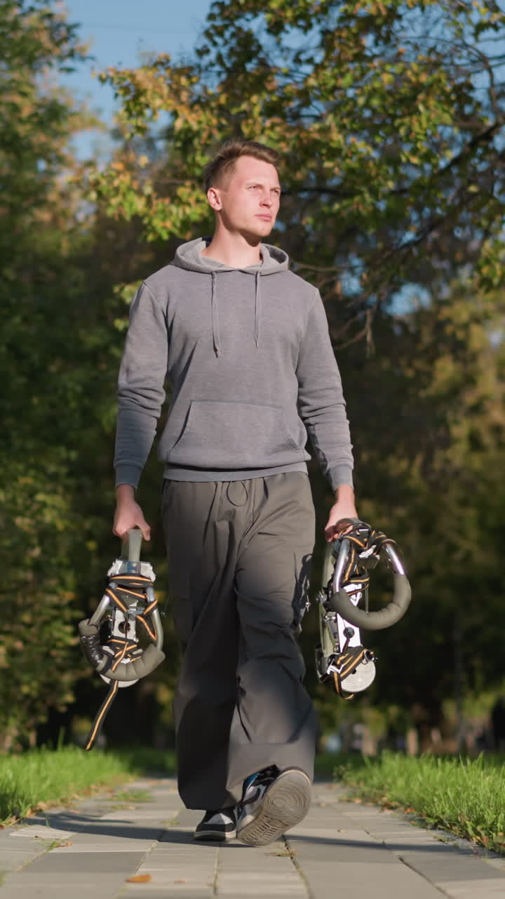 짧은 갈색 머리카락을 가진 젊은 남자가 은 자연 환경에서 푸른 나무와 풀로 둘러싸인 밝은 회색 후디와 올리브 녹색 화물 바지를 입고 스프링 로드 된 스을 들고 포장 된 길을 고 있습니다.