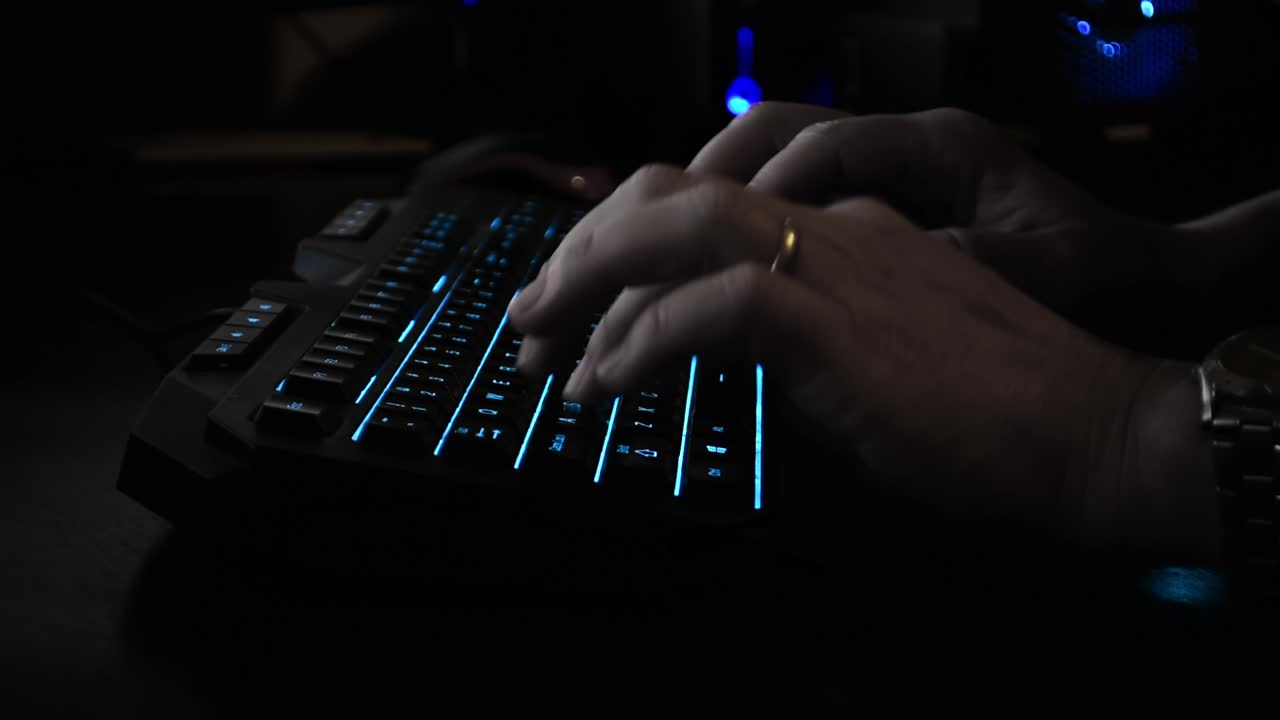 Side view of a cyber criminal's hands typing on a backlit keyboard, dark office