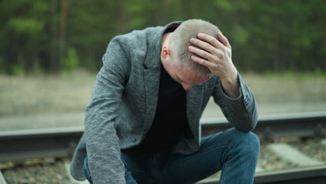 a close view of a man in a gray jacket and blue jeans, wearing canvas shoes, handling a handgun, sitting on a railway track, his head is bowed, and one hand is on his head