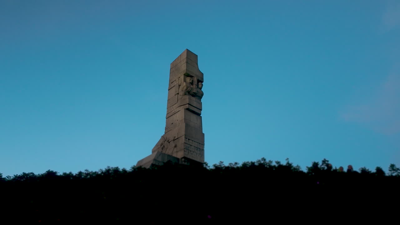 The monumental Westerplatte memorial rises on a green hill above a wide, paved viewing area and steps at twilight
