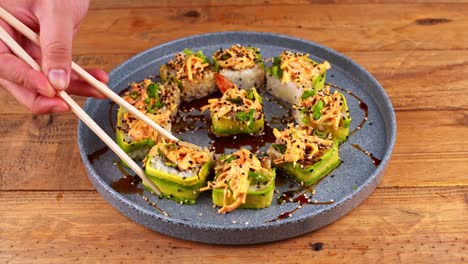 Close-up-of-hands-with-chopsticks,-awkwardly-picking-up-an-avocado-sushi-roll-from-a-blue-plate