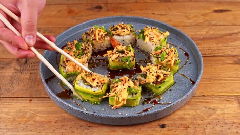 primer plano de las manos con palillos, recogiendo torpemente un rollo de sushi de aguacate de un plato azul