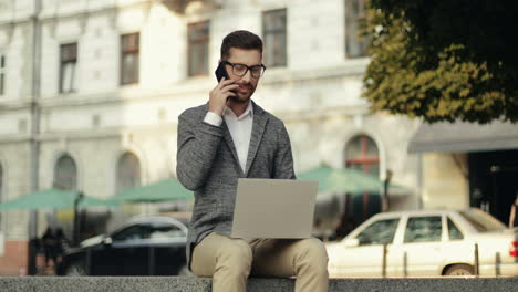 Buen-Hombre-De-Negocios-Hablando-Por-Su-Teléfono-Móvil-Mientras-Se-Sienta-En-La-Pared-De-La-Ciudad-Y-Trabaja-En-Su-Computadora-Portátil