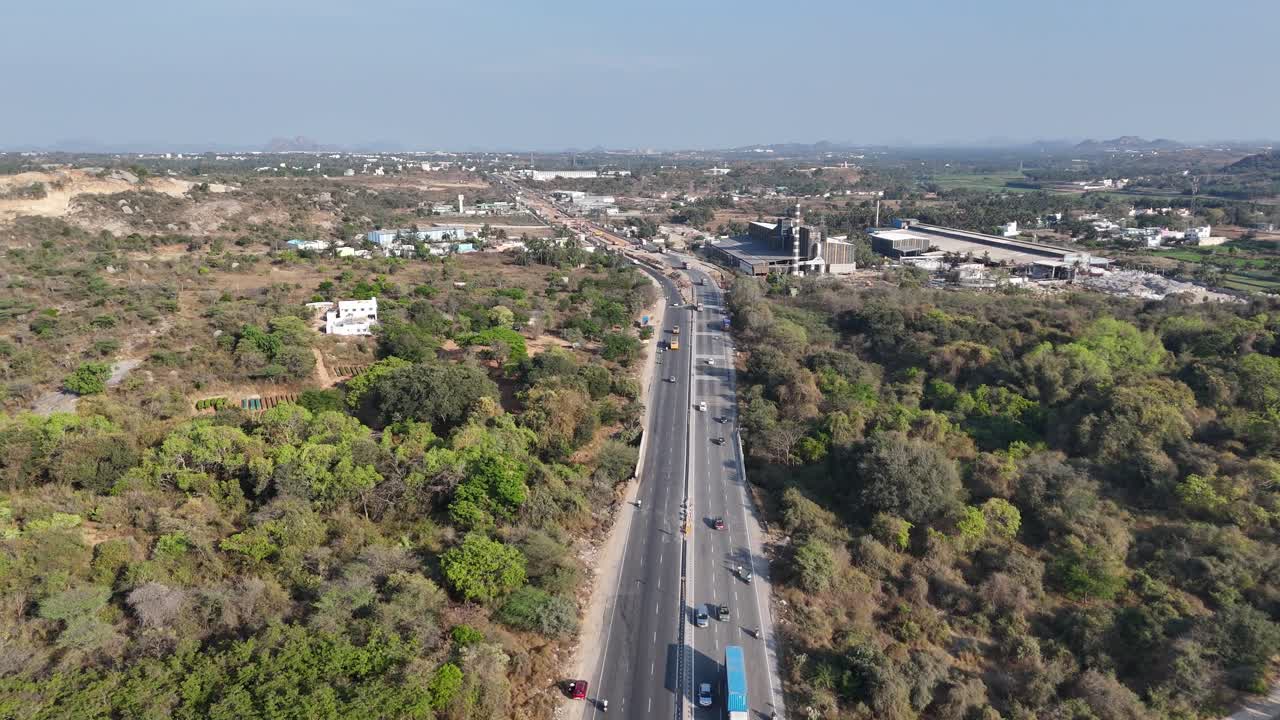An aerial long shot captures a straight, multi-lane highway stretching into the distance through a predominantly dry, shrub-covered landscape