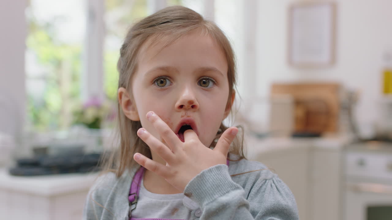 una ragazzina carina con le mani coperte di cioccolato che lecca le dita divertendosi a cuocere in cucina un bambino cattivo che si gode un gustoso dolce a casa