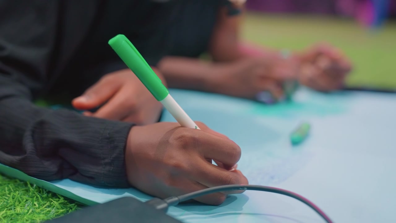 Bambino di colore che disegna su carta blu, primo piano di mani concentrate che tengono un pennarello verde su un tappeto da gioco morbido, studente che pratica la scrittura e la coordinazione motoria fine, atmosfera di apprendimento tranquilla al chiuso.
