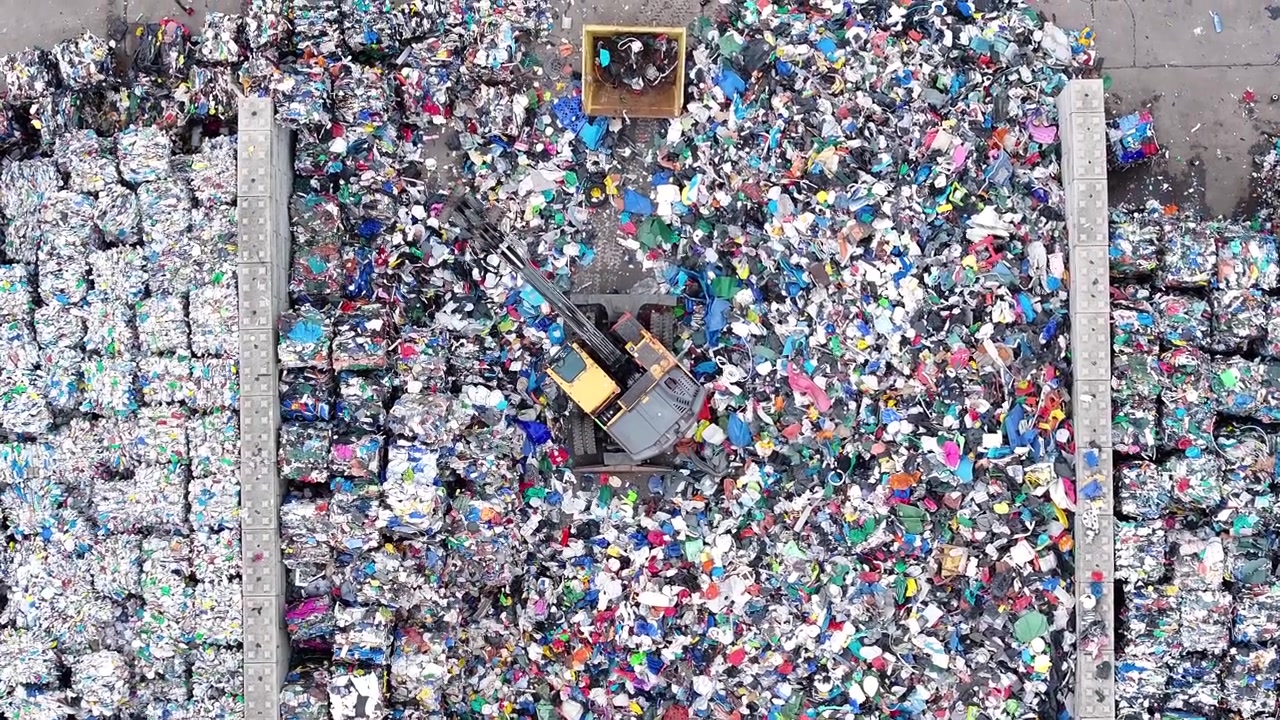 Machines sort and process plastic waste in a recycling facility. Large piles of mixed recyclable materials are visible. Sorting equipment operates to separate different plastics.