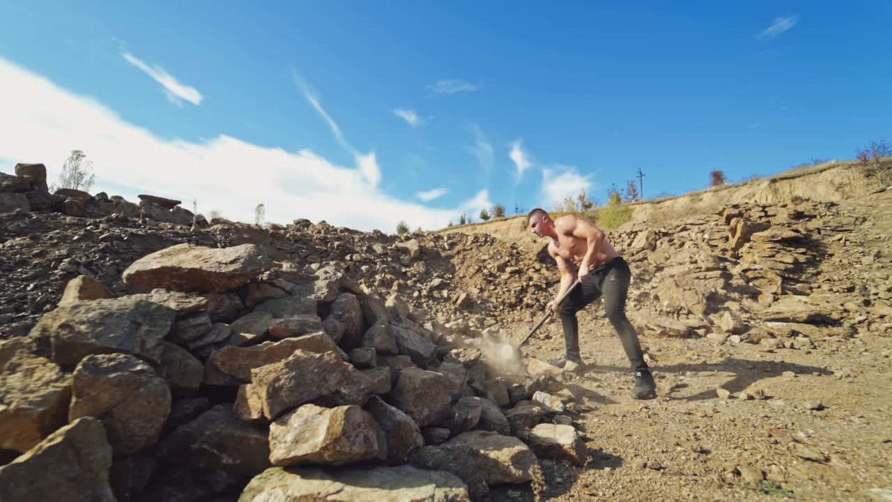 Sportsman with hard hammer on the rocky background. Athletic man without shirt breaking stones with metal hammer outdoors. Slow motion.