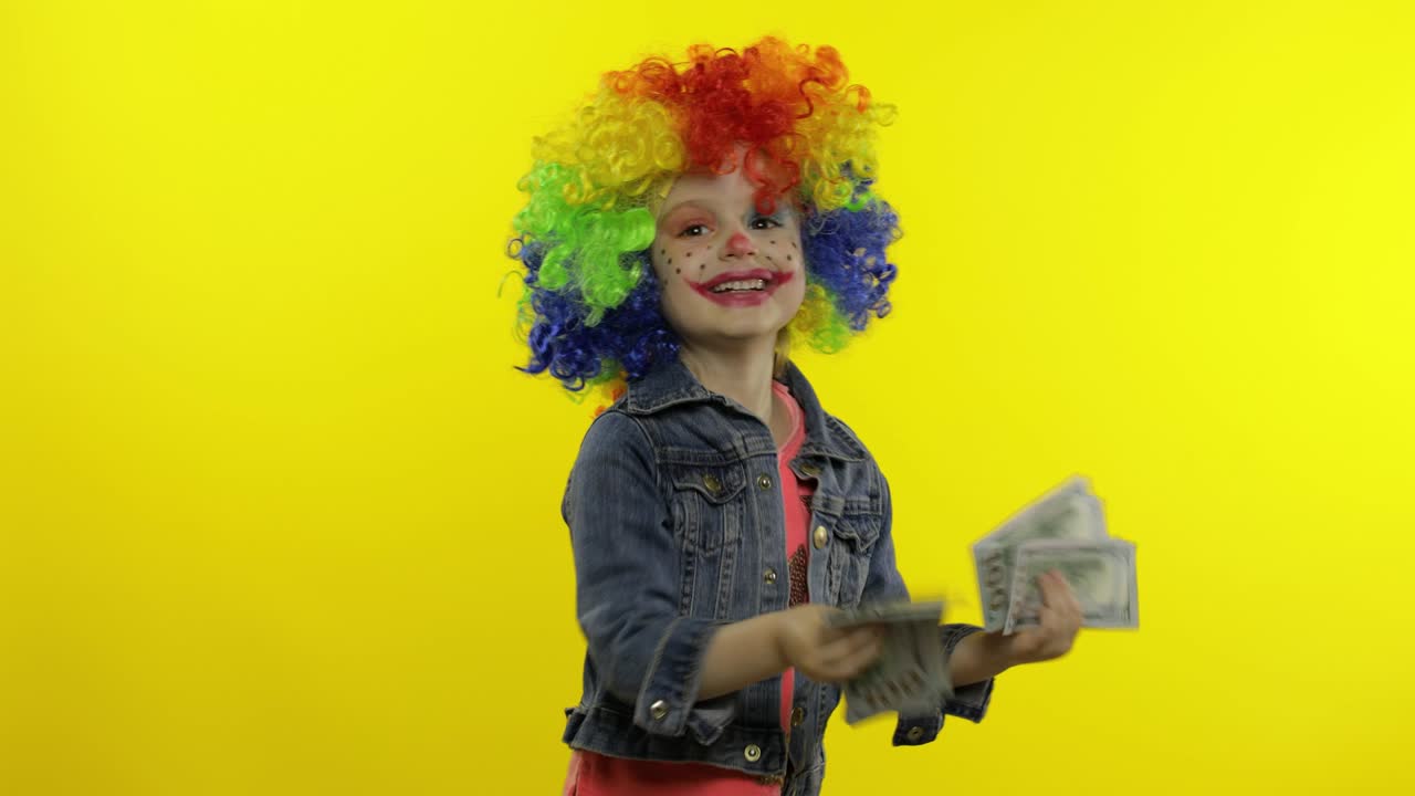 niña payaso con una peluca colorida bailando con billetes en efectivo de dólares. divirtiéndose, sonriendo