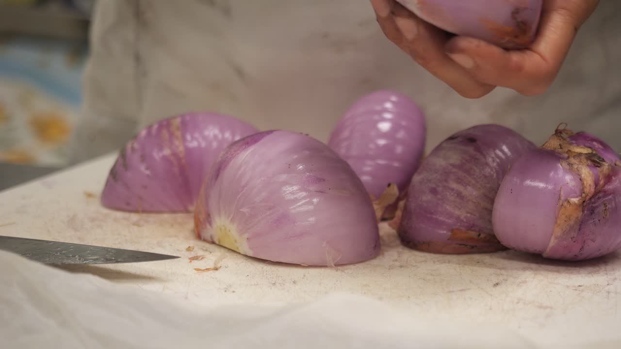 Halved red onions rest on a white cutting board beside a knife, with a chef’s hands in frame. The kitchen setting includes a blurred red-checkered background, evoking a warm prep atmosphere