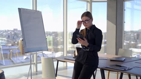 beautiful young business woman indoors. pretty woman in black clothes putting on her glasses and looking into her phone while standing in office room with panoramic windows.