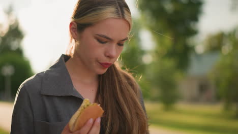 primer plano de una mujer con una camiseta gris sosteniendo shawarma, observándola pensativamente, la luz del sol suave ilumina su mano y el bocadillo, con un fondo borroso de vegetación y cálidos rayos de sol
