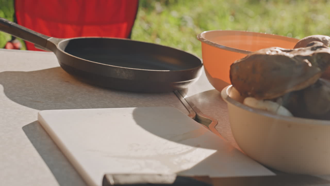 Poêle sur une table de camping avec des champignons, gros plan d'un bol, d'une planche et de la poêle sous une lumière chaude. Mains retournant la poêle, détail grésillant des ingrédients et de leur texture. Chaise pliante rouge en arrière-plan et ambiance rustique.