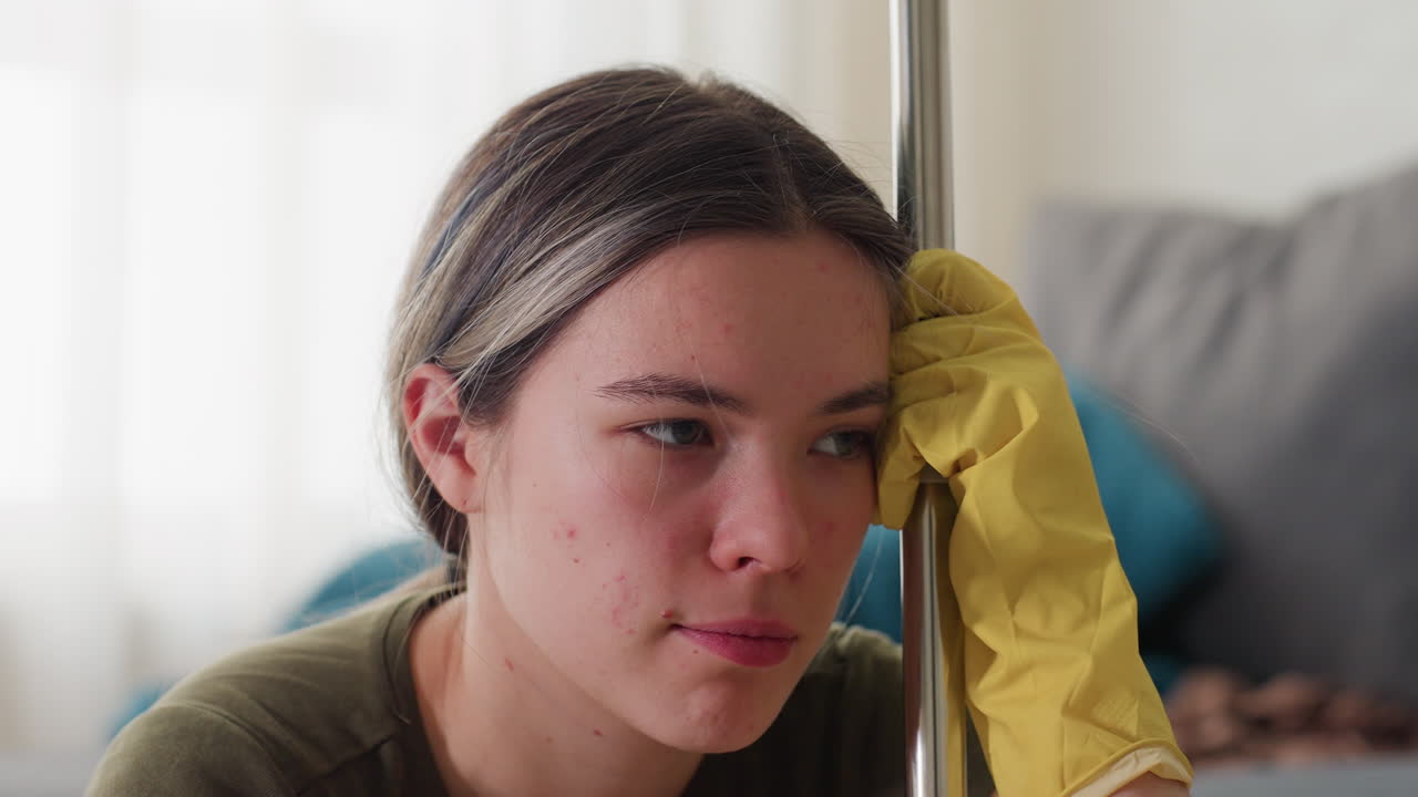 Close up of young woman wearing yellow gloves holding mop handle with tired and frustrated expression, shaking head in exhaustion after household chores, sitting in cluttered room