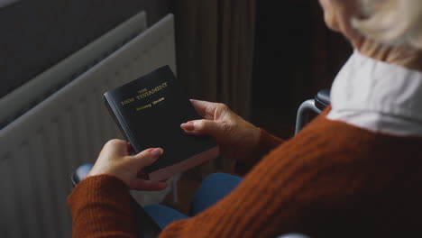 senior woman in wheelchair with bible keeping warm by radiator at home in cost of living crisis