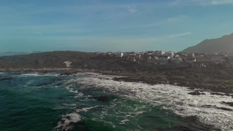 aerial-shot-moving-diagonally-while-rising-off-beach-shore-displaying-waves-and-tides-crashing-onto-rocks-as-the-view-pans-to-show-beautiful-coastal-town-Vermont-with-houses-overlooking-the-ocean