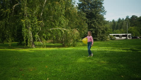 little girl walking across open grassy park area holding yellow balloon under arm while playing, with distant view of parked white buses and vehicles near forest background on sunny day