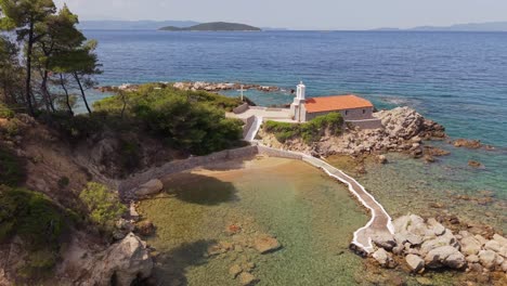 drone footage of a serene greek island in summer, featuring a small white chapel built on a rocky islet surrounded by clear blue waters and coastal beauty
