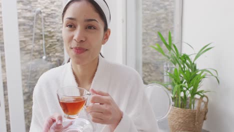 Biracial-woman-drinking-tea-and-smiling-in-bathroom