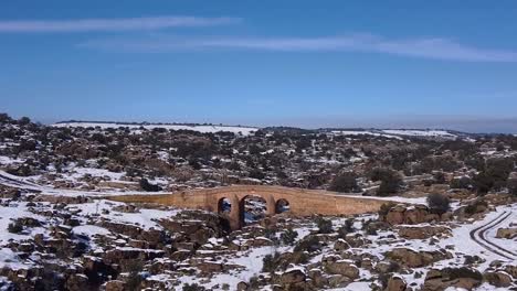 Aerial-view-of-a-drone-flying-over-a-beautiful-sunny-snowy-field-with-a-river-and-an-old-bridge