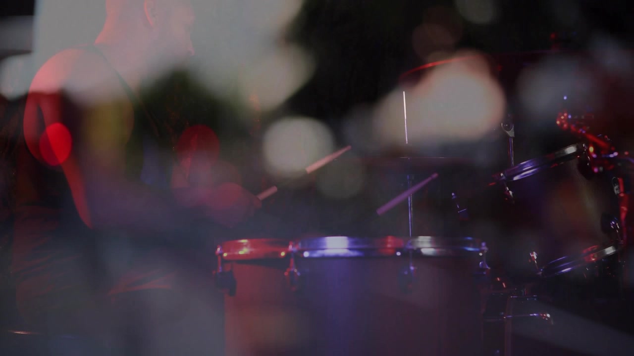 Luci del palco in movimento, telecamera bassa che sposta la messa a fuoco, rivelando una batteria con bokeh, evidenziando la musica.