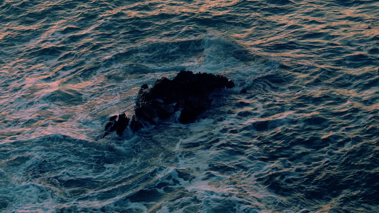 Waves Crashing on Lone Rock in Ocean with Orange Sunset Lighting Reflecting on Sea. Slow Motion Nature Swirling Sea Footage.