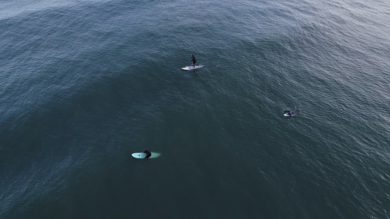 drone volando sobre el mar, dos surfistas esperando olas y una paleta de pie