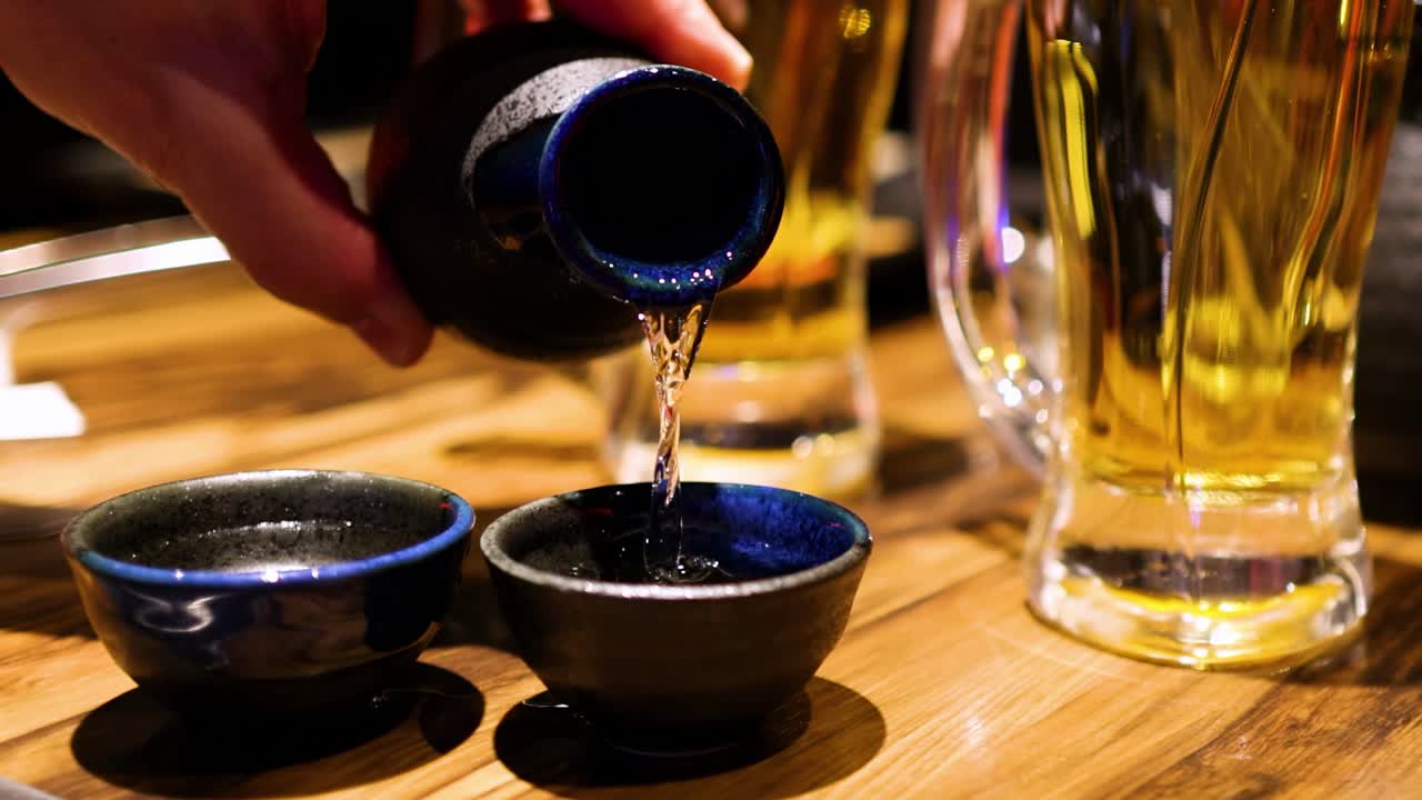 A hand pours sake into two blue ceramic cups beside tall beer glasses on a wooden table.