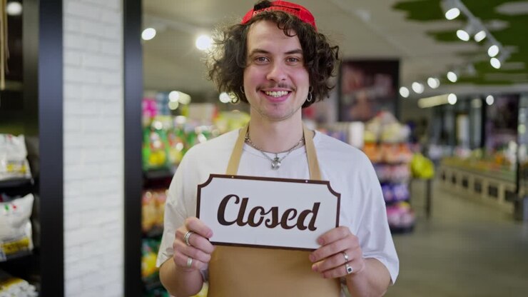 A smiling guy with curly hair and yellow front stands in a supermarket and holds a sign in front of his chest that says Closed