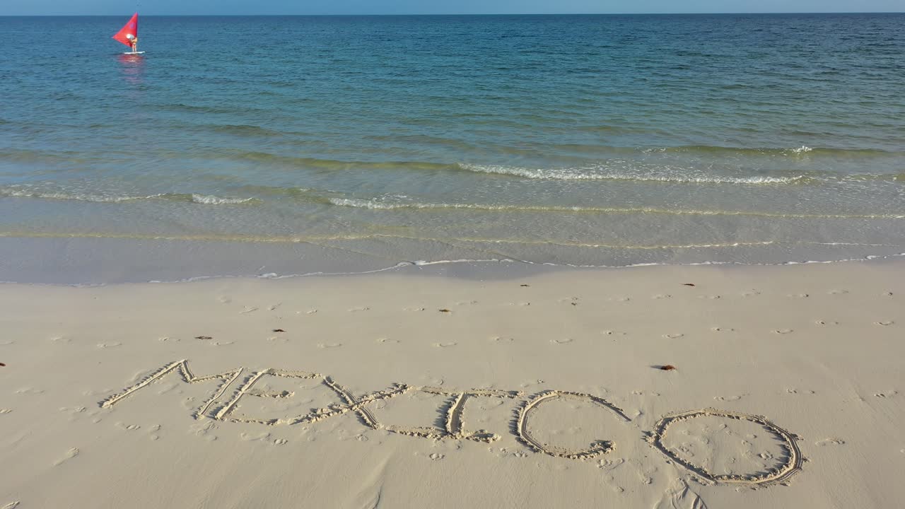 поворотный снимок мексики, вписанной в песок на пляже с виндсерфером вдалеке