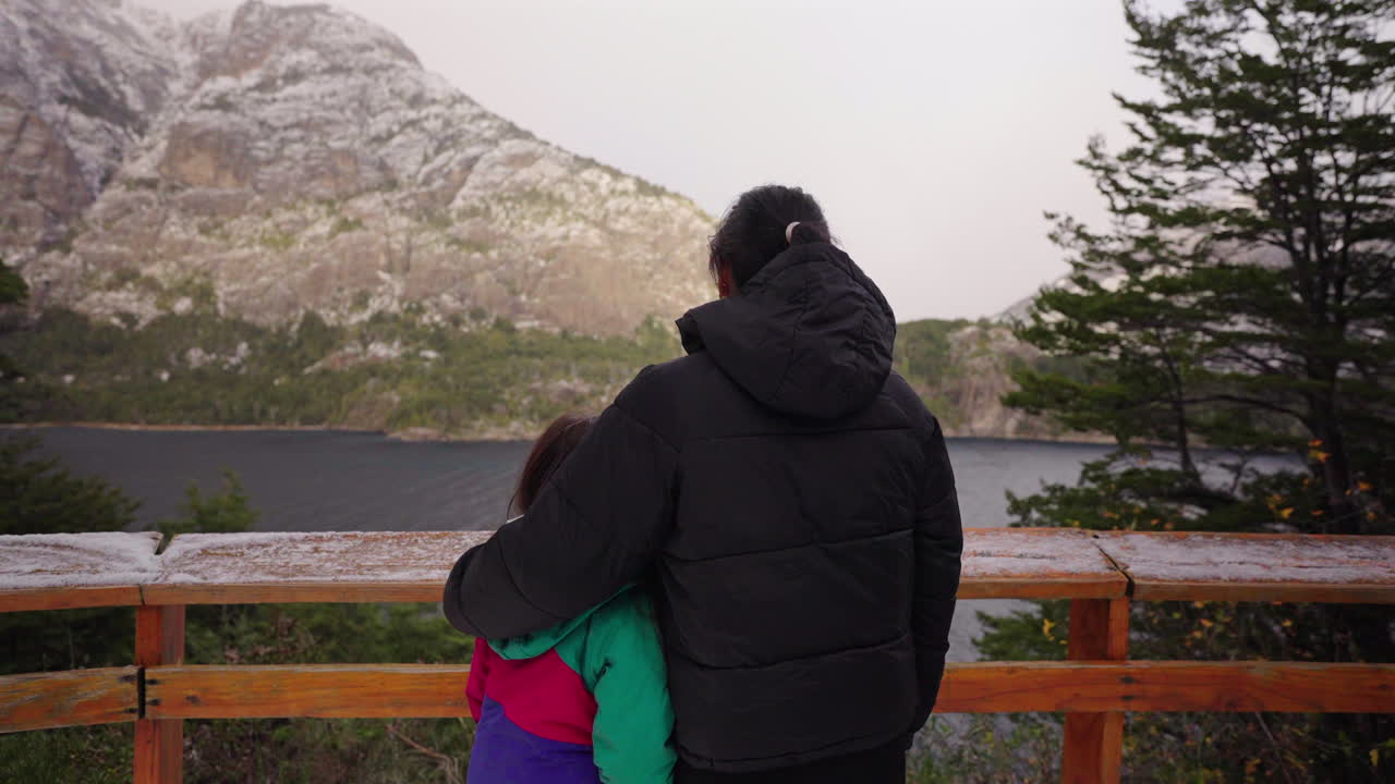 Camera slowly slides as adult and child embrace at a scenic wooden viewpoint over a lake in snowy Bariloche, Argentina. Snow-dusted mountains and pine trees complete this touching winter moment