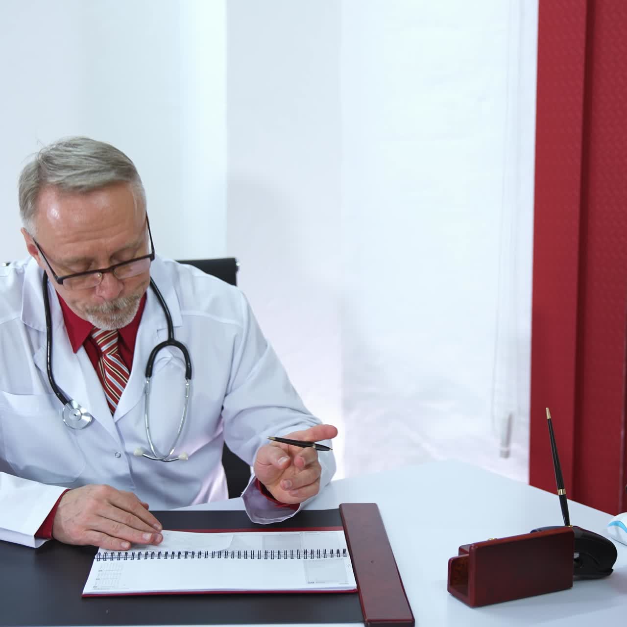 Professional medic sits at table and speaks to a patient. Senior doctor in glasses and grey beard. Phonendoscope on the neck of a phisician