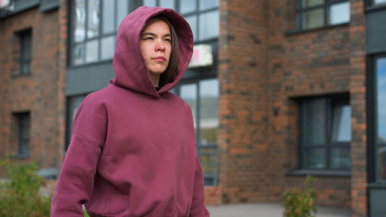 Active woman in wine hoodie lifts dumbbells outdoors in courtyard, scanning surroundings during strength session, showing focus, discipline, healthy lifestyle motivation amid urban greenery