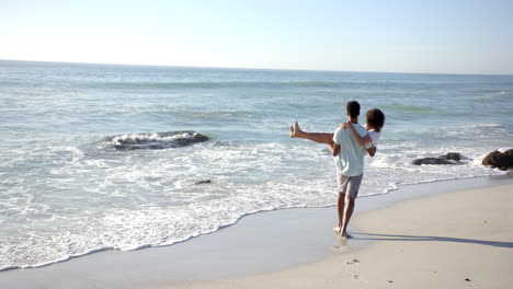 Una-Pareja-Birracial-Disfruta-De-Un-Día-De-Playa-Con-Espacio-Para-Copiar,-Con-El-Hombre-Cargando-A-La-Mujer-A-Cuestas