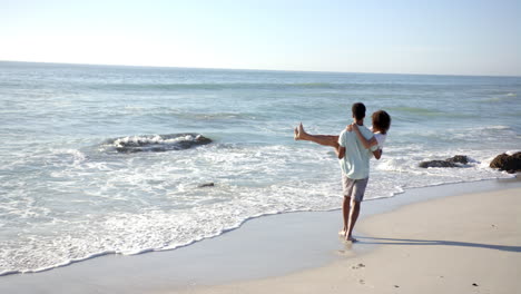 una pareja biracial disfruta de un día en la playa con espacio de copia, con el hombre llevando a la mujer a la espalda