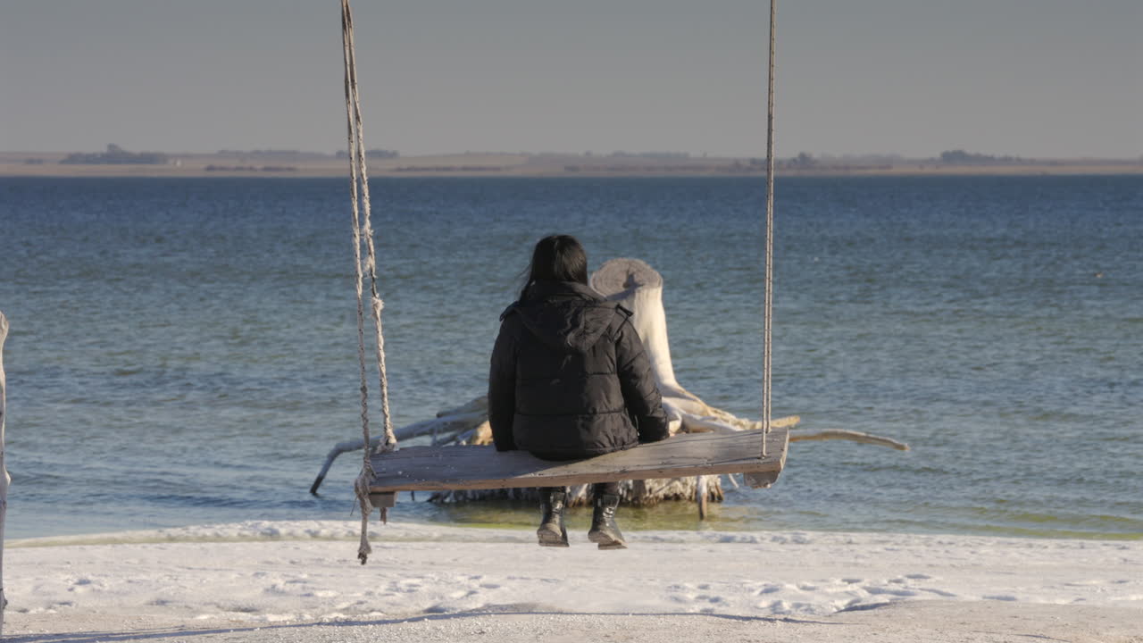 Giovane donna su una grande altalena di legno di fronte a un lago salato calmo a Villa Epecuen, Argentina. La scena presenta una radice d'albero sbiadita dal sale e un'atmosfera pomeridiana dai toni seppia. Inquadratura statica generale.